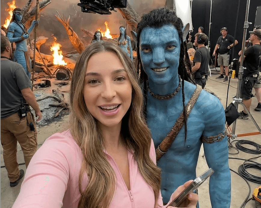 Girl in pink shirt standing next to Jake Sully in Avatar form taking a selfie behind the scenes on the movie set